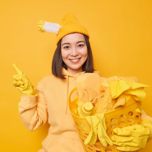 Pleased Asian woman does laundry at home wears hat with stuck toilet brush hoodie and rubber gloves points away on blank copy space isolated over yellow background shows product for cleaning