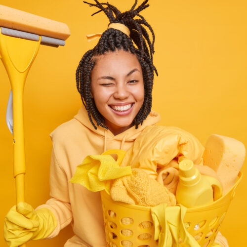 Positive Afro American housewife winks eye smiles broadly has happy mood after cleaning house holds mop for washing floor carries laundry basket isolated over yellow background removes mess.