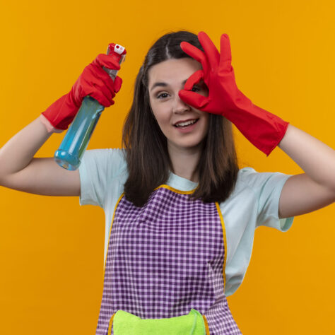 young beautiful girl in apron and rubber gloves holding cleaning spray smiling cheerfully doing ok sign looking through this sign standing over orange background