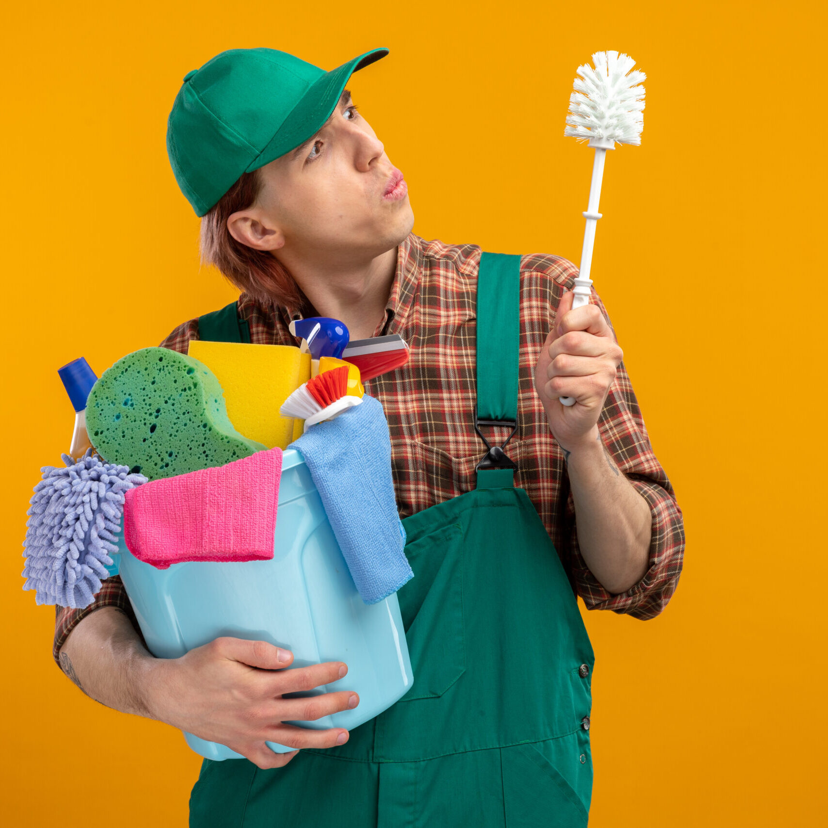 young cleaning man in plaid shirt jumpsuit and cap holding cleaning brush and bucket with cleaning tools looking at brush intrigued standing over orange background